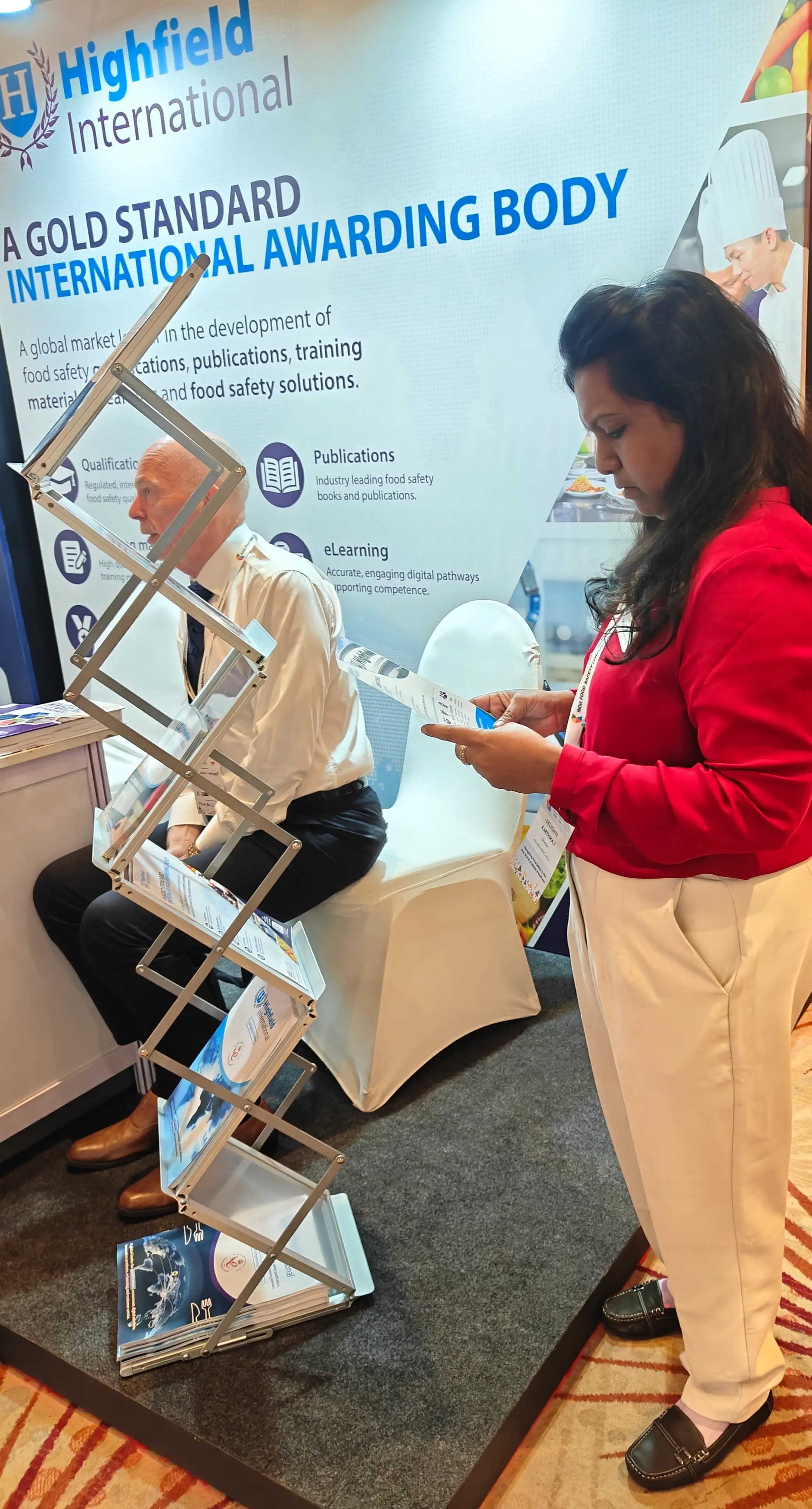A delegate speaking with a visitor beside a roll-up banner and brochure stand at an exhibition.