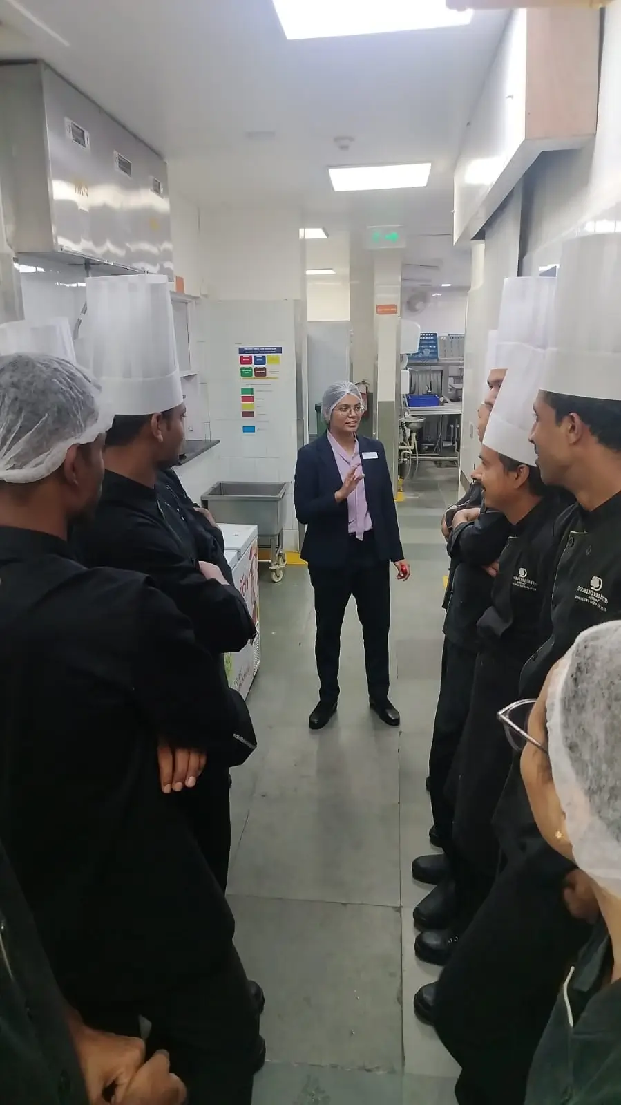Kitchen staff wearing hairnets gathered in a circle for a briefing inside a food preparation area.