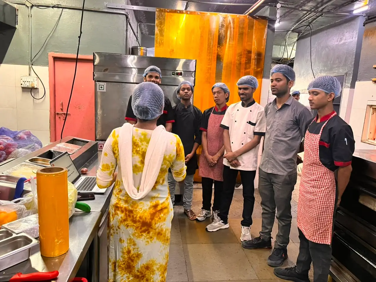 Kitchen staff in hairnets and aprons standing in a line listening to a trainer inside a commercial kitchen.