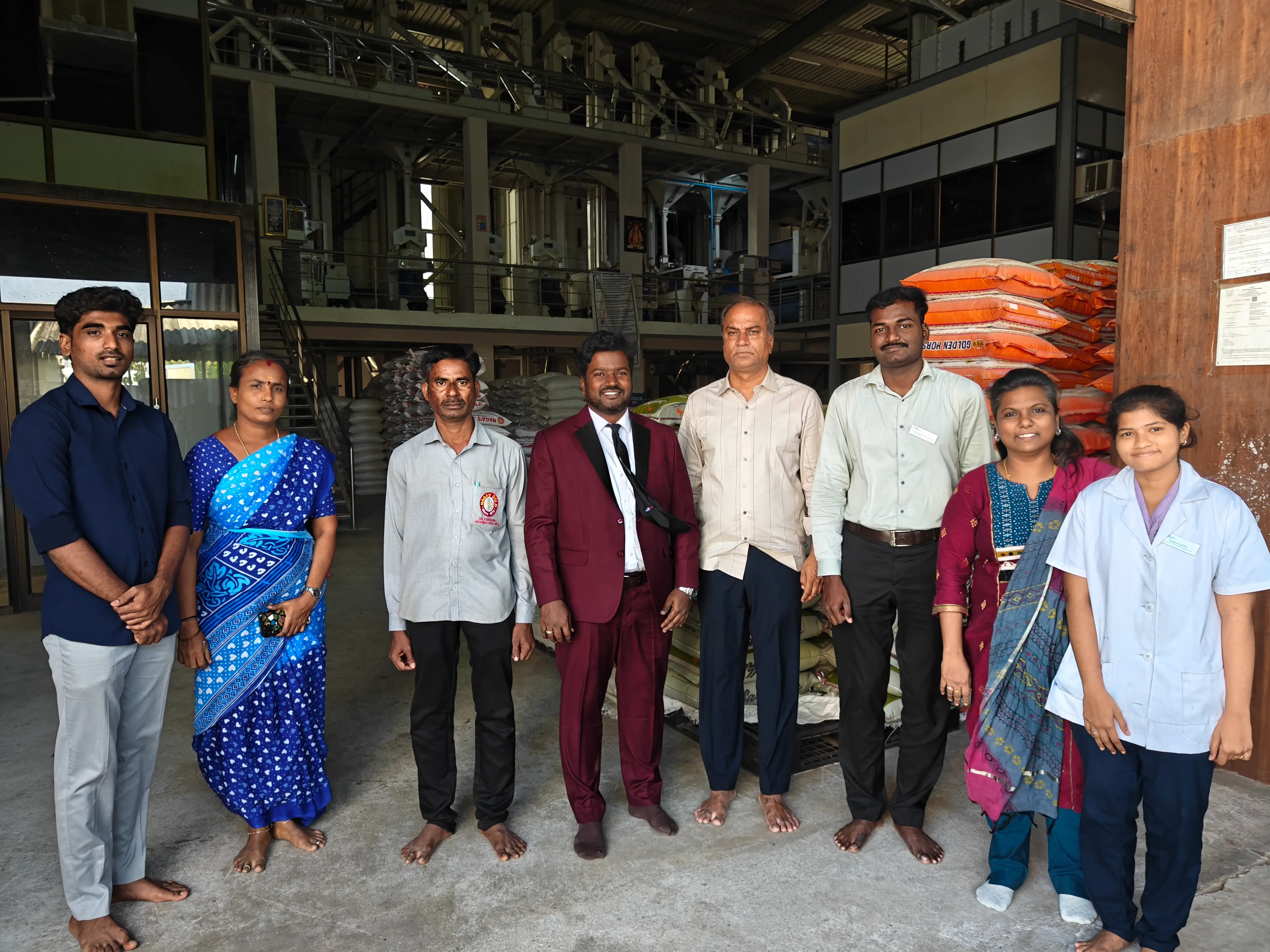 Group of food processing workers and visiting professionals standing together inside a grain milling facility with stacked sacks of produce.