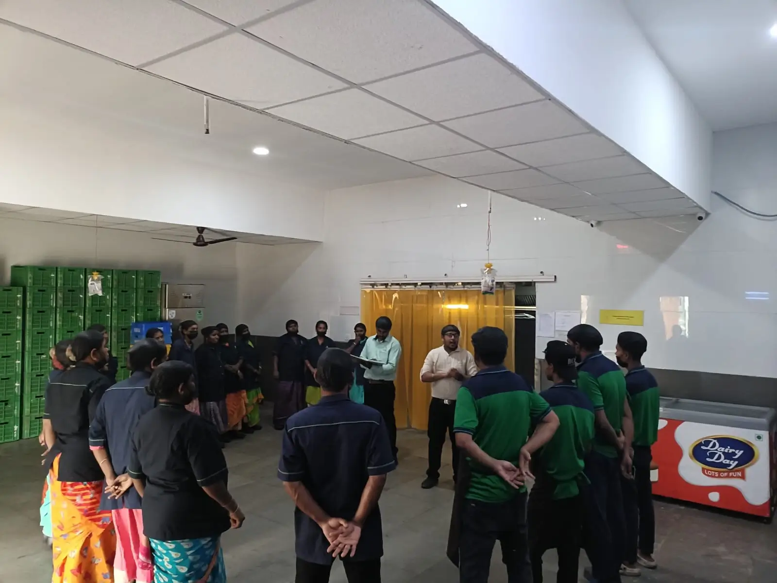 Warehouse staff standing in a circle for a briefing beside stacked crates and a cold storage door.