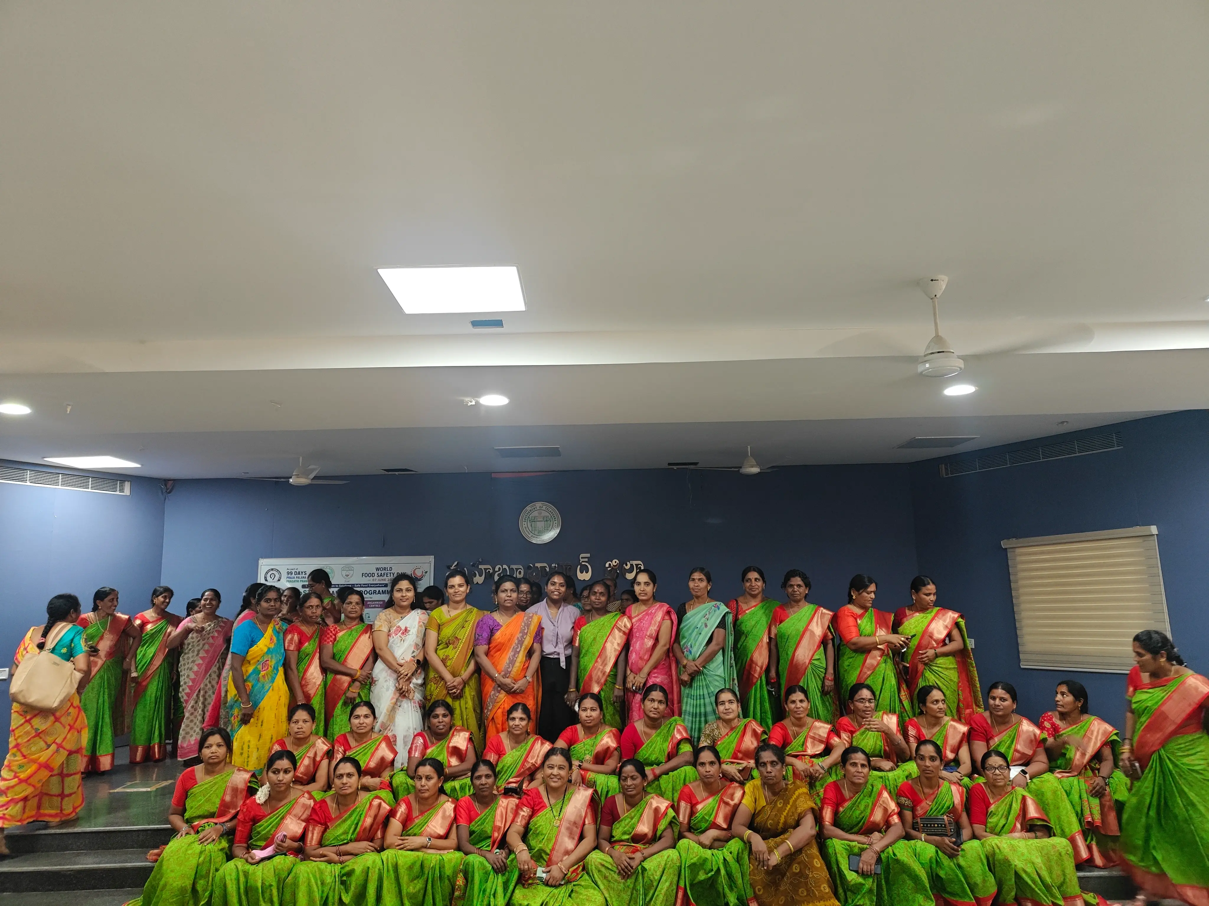 A large group of women in matching green and red sarees seated and standing together in a training hall.